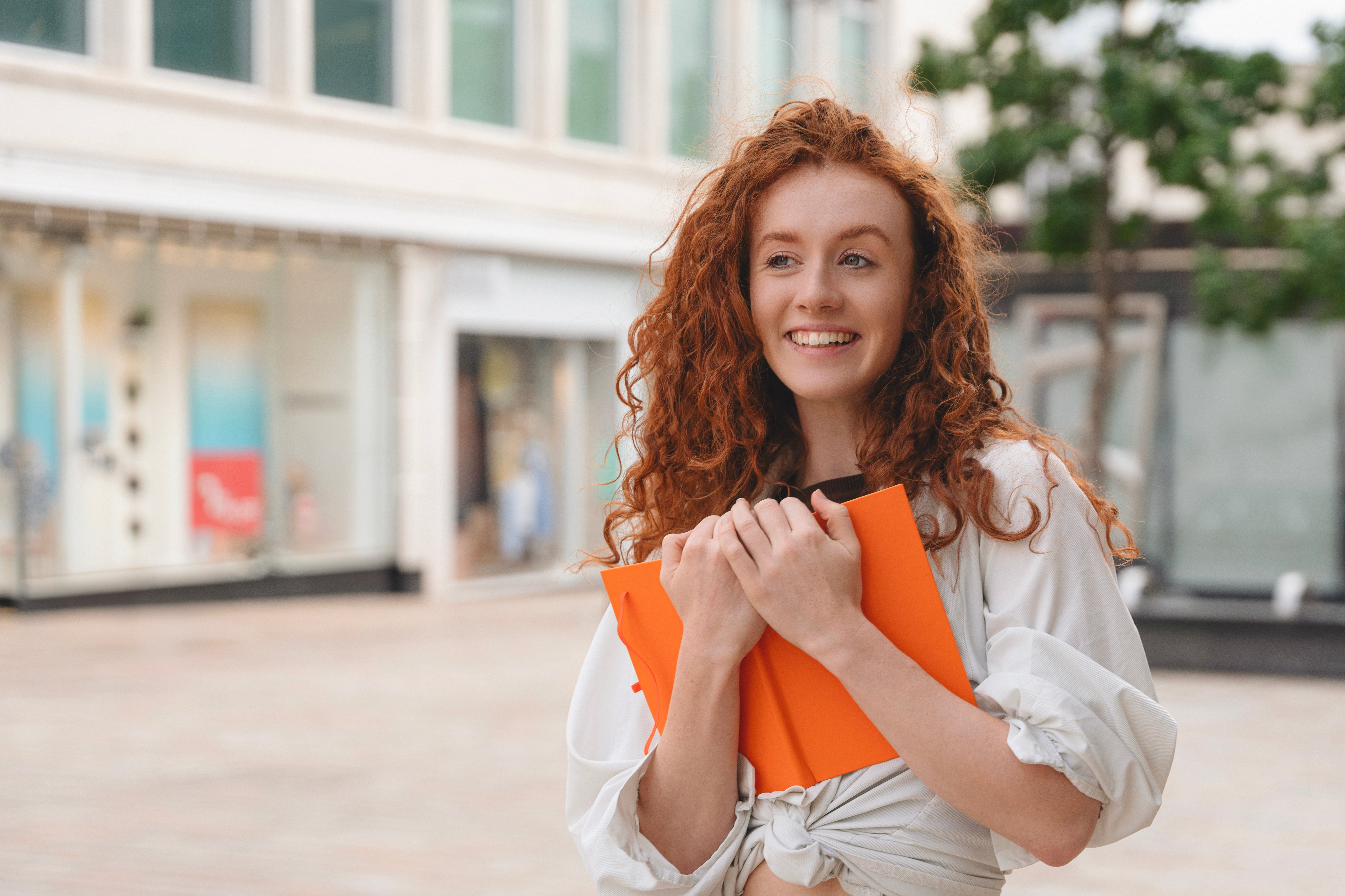 A cheerful young woman stands outdoors in a bustling city, holding bright orange folders close to her chest with a joyful expression.
